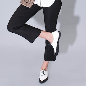 May include: A pair of white leather lace-up shoes with black trim and laces. The shoes are worn by a person wearing black pants and a white shirt.
