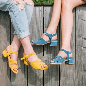May include: Two people wearing sandals with contrasting colors. One person is wearing yellow sandals and the other is wearing blue sandals. Both sandals have a strap across the ankle and a thick heel.
