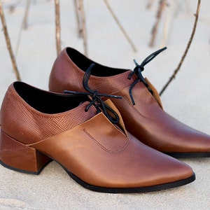 Op de afbeelding: Een paar bruine leren schoenen met hak en zwarte veters. De schoenen hebben een spitse neus en een dikke hak.