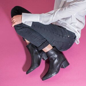 以下が含まれることがあります： 黒のジーンズと白いボタンダウンシャツを着た人が、足を組んで座っています。彼らは、尖ったつま先の黒いアンクルブーツを履いています。
