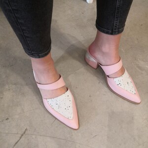 May include: A pair of pink and white heeled mules with a pointed toe and a black strap across the top. The shoes have a speckled white design on the pink portion.