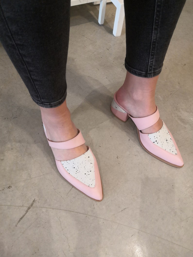 May include: A pair of pink and white heeled mules with a pointed toe and a black strap across the top. The shoes have a speckled white design on the pink portion.