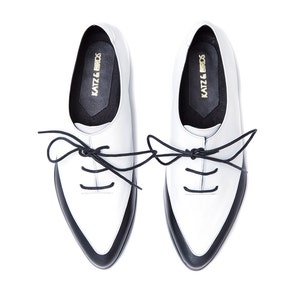 May include: A pair of white leather lace-up shoes with black trim and black laces. The shoes have a pointed toe and a low heel. The brand name "KATZ & BIRDS" is printed on the inside of each shoe.