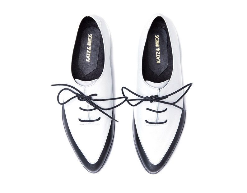 May include: A pair of white leather lace-up shoes with black trim and black laces. The shoes have a pointed toe and a low heel. The brand name "KATZ & BIRDS" is printed on the inside of each shoe.