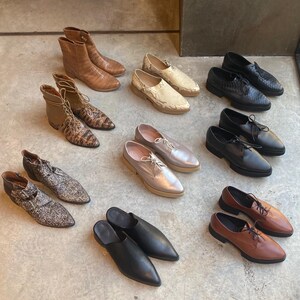 May include: A collection of nine different styles of shoes in various colors and materials, including black leather, brown leather, snakeskin, and silver. The shoes are arranged on a light gray concrete floor.