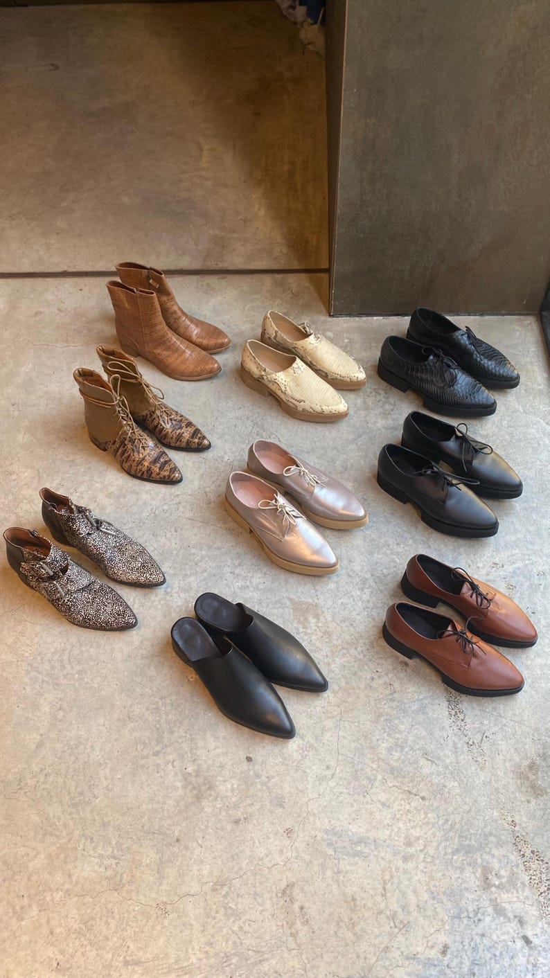May include: A collection of nine different styles of shoes in various colors and materials, including black leather, brown leather, snakeskin, and silver. The shoes are arranged on a light gray concrete floor.