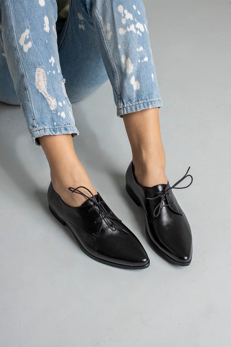 May include: A pair of black leather lace-up shoes with pointed toes. The shoes are worn by a person wearing blue jeans.