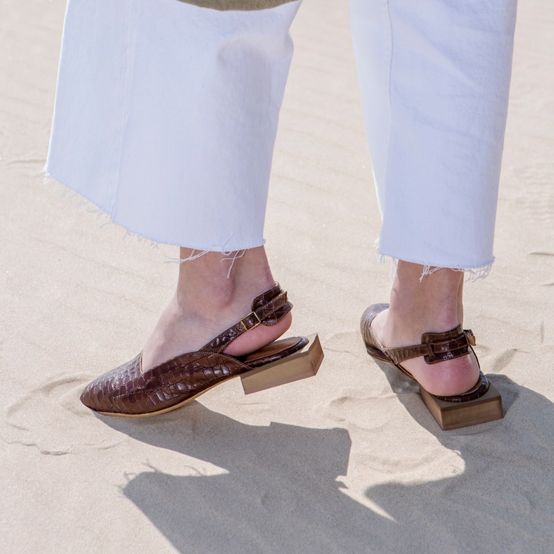 Op de afbeelding: Een paar bruine leren sandalen met hak en een gesp. De sandalen hebben een gestructureerd leren bovenwerk en een blokhak.