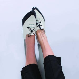 May include: A pair of white and black leather shoes with black laces. The shoes are worn by a person with black pants.