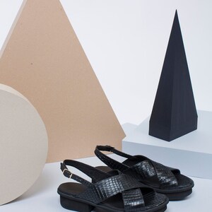K&ouml;nnte beinhalten: Ein Paar schwarze Ledersandalen mit einem Kreuzriemen-Design und einem dicken Absatz.