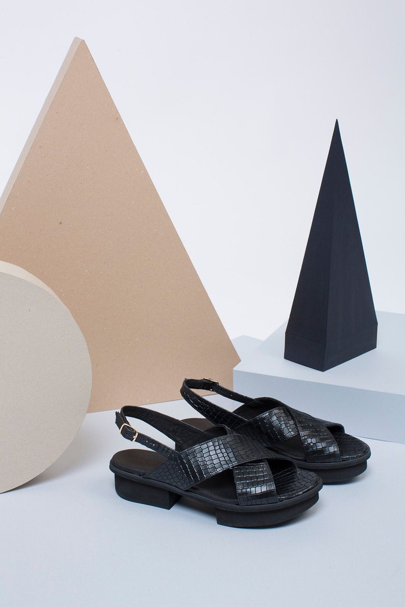 K&ouml;nnte beinhalten: Ein Paar schwarze Ledersandalen mit einem Kreuzriemen-Design und einem dicken Absatz.