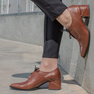 Op de afbeelding: Bruine leren veterschoenen met een dikke hak. De schoenen hebben een spitse neus en worden gedragen door iemand met een zwarte broek.