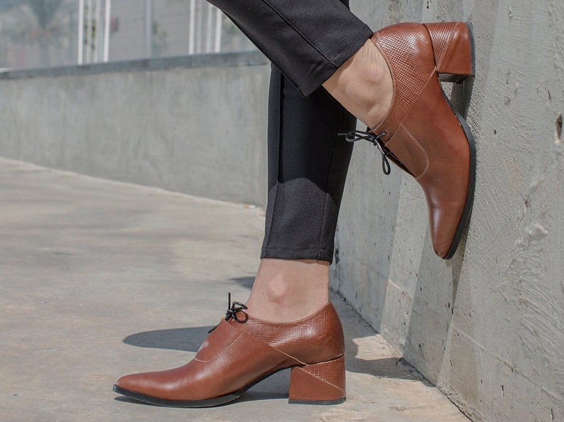 Op de afbeelding: Bruine leren veterschoenen met een dikke hak. De schoenen hebben een spitse neus en worden gedragen door iemand met een zwarte broek.