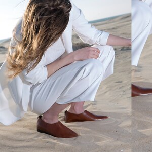 May include: A woman wearing a white blazer and pants sits on a sandy beach. She is wearing brown leather slip-on shoes with pointed toes.