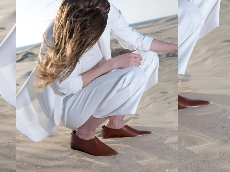 May include: A woman wearing a white blazer and pants sits on a sandy beach. She is wearing brown leather slip-on shoes with pointed toes.