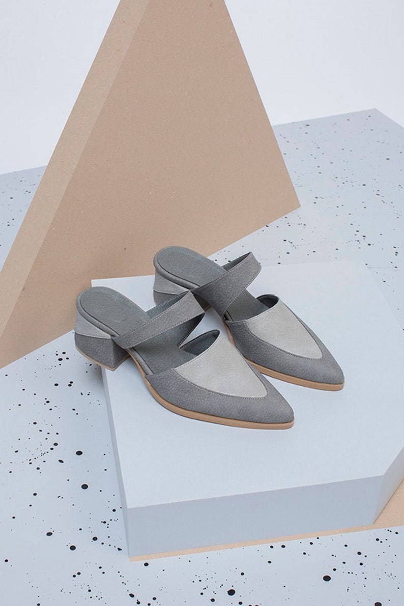 May include: A pair of gray suede heeled mules with a pointed toe and a strap across the top of the foot. The shoes are on a white platform.