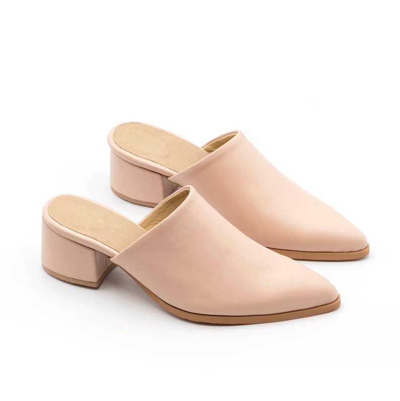 May include: A pair of light pink heeled mules with a pointed toe. The mules have a block heel and are made of a smooth leather-like material.