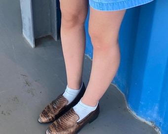 May include: Brown and tan snakeskin-patterned loafers with a dark brown sole. The shoes are worn with white socks and blue denim shorts. The background includes a blue wall and a gray floor.