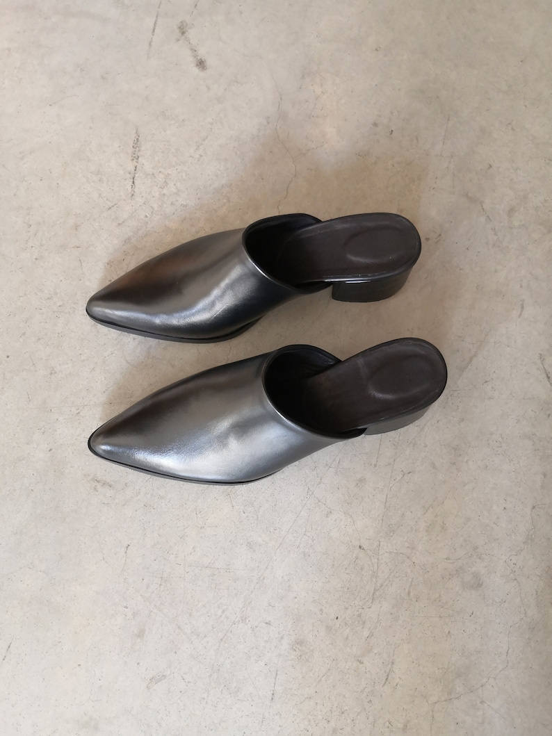 May include: A pair of black leather mules with pointed toes and small block heels. The shoes have a sleek, minimalist design, and are set against a neutral background.
