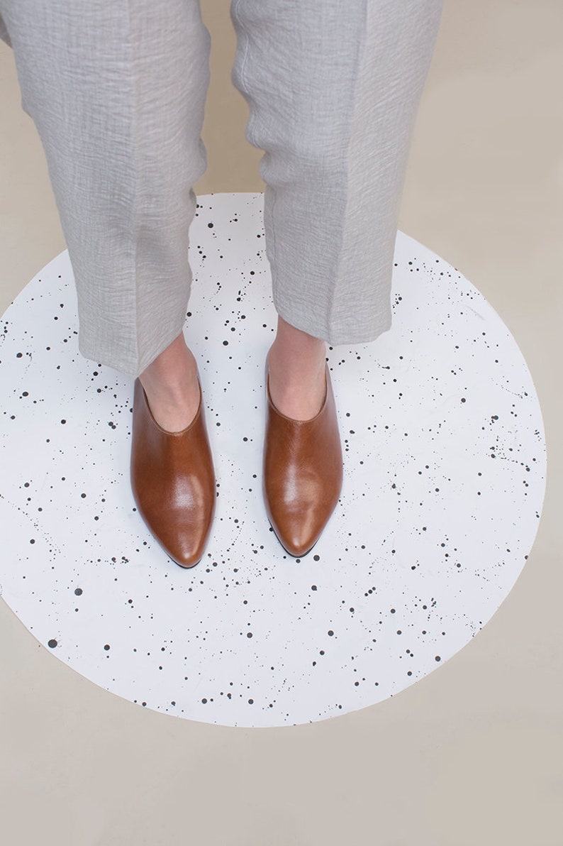 May include: A pair of brown leather heeled mules with pointed toes on a white background with black splatter paint.