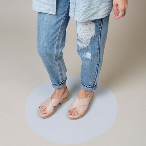 K&ouml;nnte beinhalten: Ein Paar hellrosa Ledersandalen mit gekreuzten Riemen und einem niedrigen Absatz. Die Sandalen werden von einer Person getragen, die eine blaue Jeans tr&auml;gt.