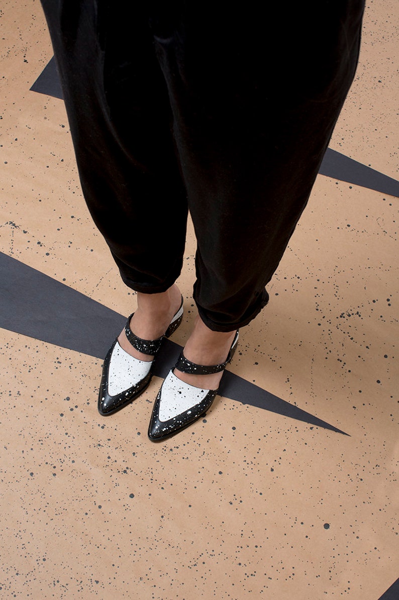 May include: A pair of black and white leather shoes with a pointed toe and a strap across the top. The shoes have a splatter paint design.