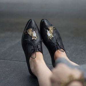 Puede incluir: Un par de zapatos oxford de cuero negro con adornos de cuentas doradas y negras en forma de p&aacute;jaros. Los zapatos tienen cordones y se llevan puestos.