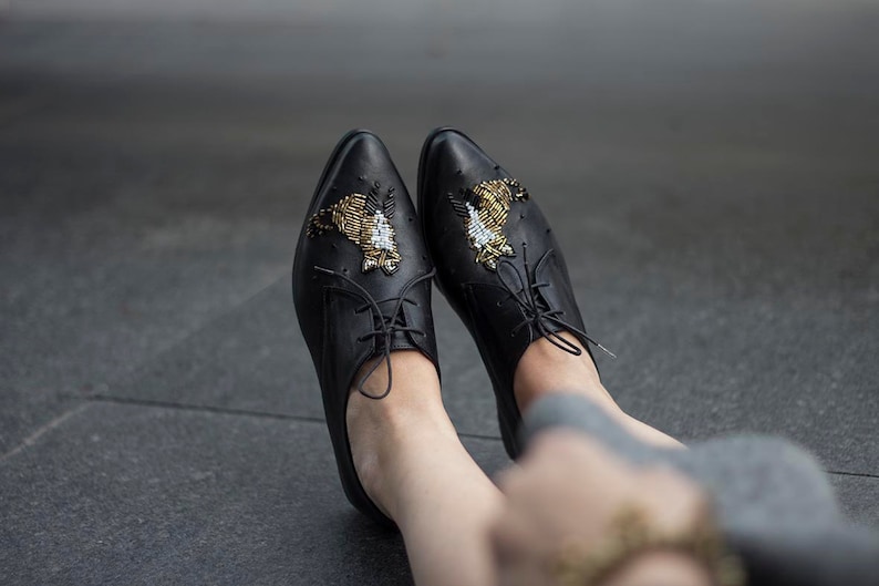 Puede incluir: Un par de zapatos oxford de cuero negro con adornos de cuentas doradas y negras en forma de p&aacute;jaros. Los zapatos tienen cordones y se llevan puestos.