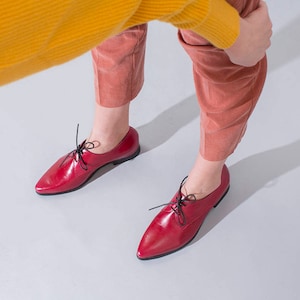 May include: A pair of red leather Oxford shoes with black laces. The shoes have a pointed toe and a low heel. The shoes are shown from a high-angle perspective.