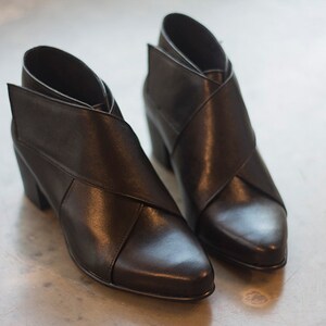 May include: A pair of black leather ankle boots with a distinctive crisscross design. The boots feature a pointed toe and a block heel. The heel height is approximately 7.6 cm.