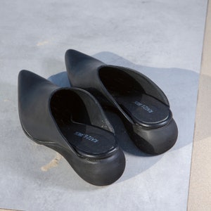 May include: A pair of black leather pointed-toe heeled mules with a thick rubber sole. The shoes have the brand name "KAIZER WDC" printed on the insole.