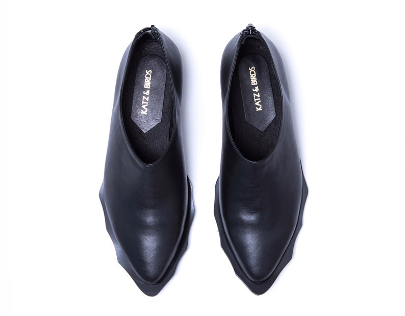 May include: A pair of black leather shoes with a pointed toe and a unique, jagged sole. The shoes have a slip-on design and feature the text "KATZ & BIRDS" on the inside.