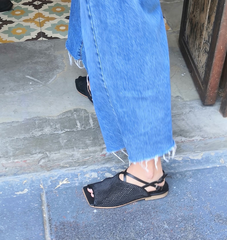 May include: Black mesh sandals with a strap across the foot and a peep toe. The sandals are worn with blue denim jeans.