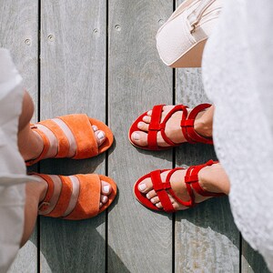 K&ouml;nnte beinhalten: Zwei Paar Sandalen, ein Paar ist orange und das andere Paar ist rot. Die Sandalen befinden sich auf einem Holzboden.