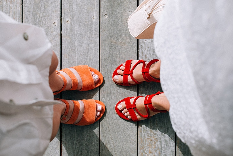 K&ouml;nnte beinhalten: Zwei Paar Sandalen, ein Paar ist orange und das andere Paar ist rot. Die Sandalen befinden sich auf einem Holzboden.