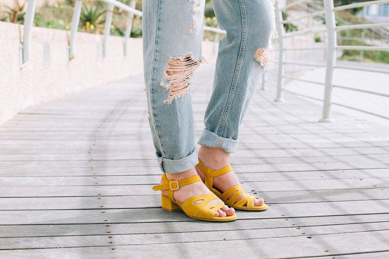 May include: A pair of yellow suede heeled sandals with ankle straps. The sandals are worn by a person wearing ripped denim jeans.