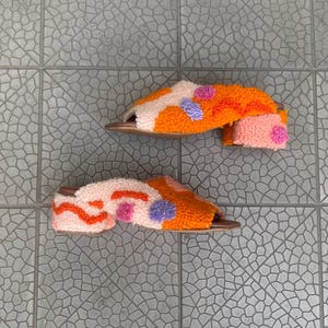 Puede incluir: Un par de sandalias de color naranja, rosa y morado con un diseño abstracto único. Las sandalias tienen un tacón bajo y están hechas de un material suave y cómodo.