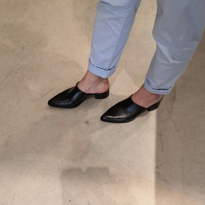 May include: Black leather mules with a pointed toe and low heel. The shoes are worn with light blue pants with rolled-up cuffs. The image is taken from a low angle, showing the shoes and the lower legs.