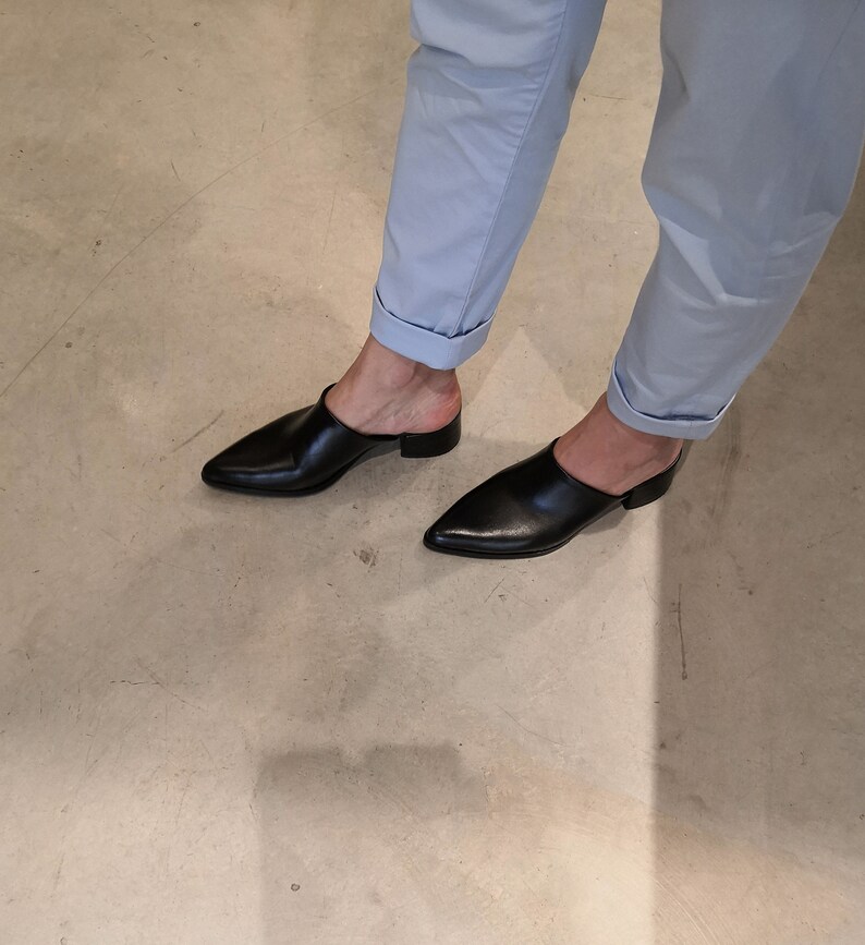 May include: Black leather mules with a pointed toe and low heel. The shoes are worn with light blue pants with rolled-up cuffs. The image is taken from a low angle, showing the shoes and the lower legs.