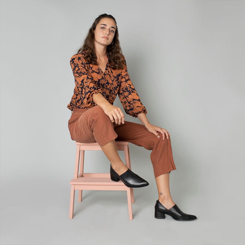 May include: A woman wearing a brown and black floral print blouse, brown pants, and black heeled shoes sits on a pink stool.