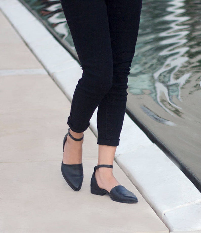 May include: A pair of black ankle strap flats with a pointed toe. The shoes are worn by a person wearing black jeans.