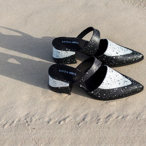 May include: A pair of black and white heeled mules with a splatter paint design. The shoes have a pointed toe and a thick block heel. The brand name "Katz & Birds" is printed on the insole.