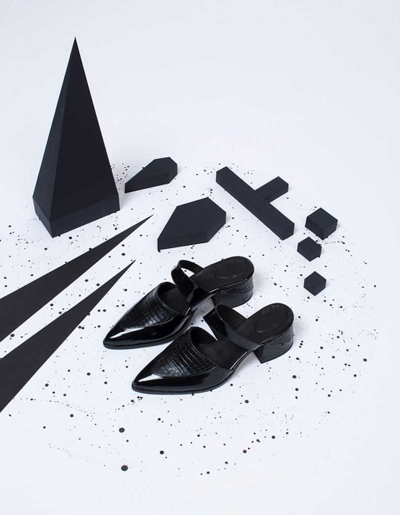 Puede incluir: Dos mules negras de tac&oacute;n de cuero con punta puntiaguda y una tira en la parte superior. Los zapatos est&aacute;n sobre una superficie blanca con formas geom&eacute;tricas negras y salpicaduras de pintura negra.