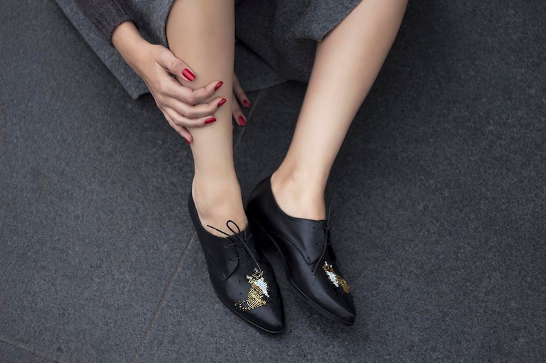 Puede incluir: Zapatos negros de cuero con cordones y adornos de cuentas doradas y blancas en los dedos.