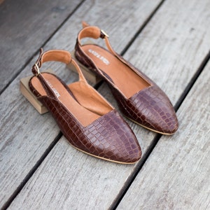 Op de afbeelding: Een paar bruine leren slingbacks met een krokodillenprint. De schoenen hebben een spitse neus en een lage hak.