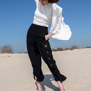 May include: A woman wearing a white top, black pants with decorative buttons, and white and black shoes. She is standing in a desert setting with sand dunes in the background.