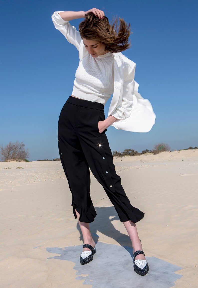 May include: A woman wearing a white top, black pants with decorative buttons, and white and black shoes. She is standing in a desert setting with sand dunes in the background.