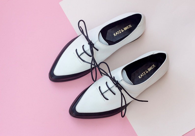 May include: Pair of white leather lace-up shoes with black trim and black laces. The shoes have a pointed toe and a low heel. The brand name "KATZ & BIRDS" is printed on the inside of each shoe.