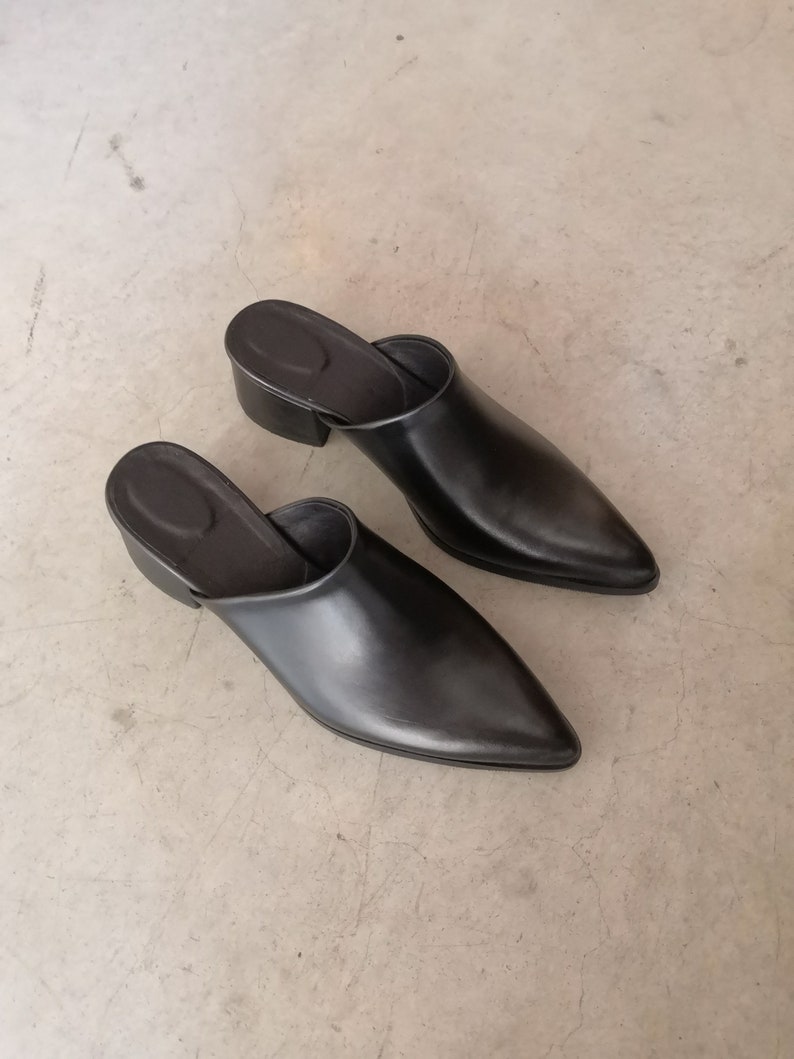 May include: A pair of black leather mules with a pointed toe and a low, block heel. The open-back design and sleek silhouette suggest a minimalist aesthetic. The shoes are set against a neutral background.