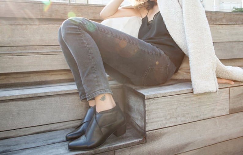 May include: Black leather ankle boots with a low heel. The boots are paired with dark grey denim jeans. A black top and a white fluffy jacket complete the outfit. The person is sitting on wooden steps.
